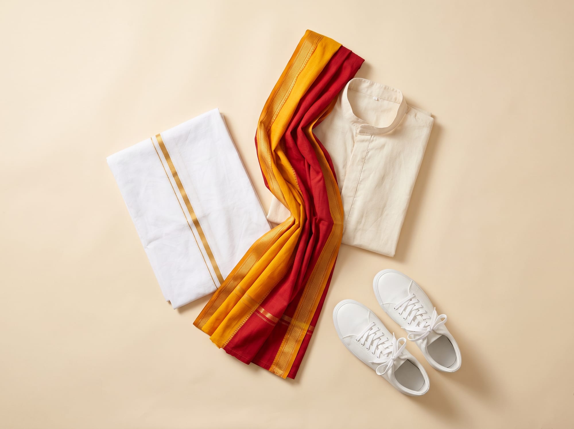 Flat lay of a white panche, a cream kurta, a red-and-yellow shalya and white sneakers.